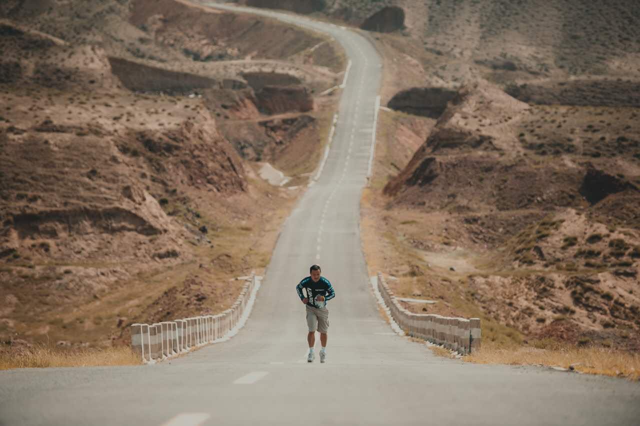 James TJ Lee running alone on a long road through a dry, hilly landscape.